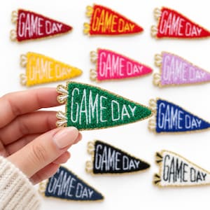 May include: A collection of triangular pennant-shaped patches in various colors including red, yellow, pink, purple, green, blue, black, and white. Each patch features the text GAME DAY embroidered in white or yellow thread.