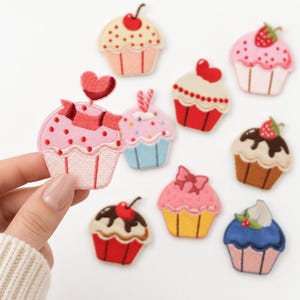 May include: A collection of embroidered cupcake patches featuring various designs, including pink frosting with a heart topper, chocolate drizzle, strawberries, cherries, and bows, rendered in vibrant colors like red, blue, yellow, and brown.