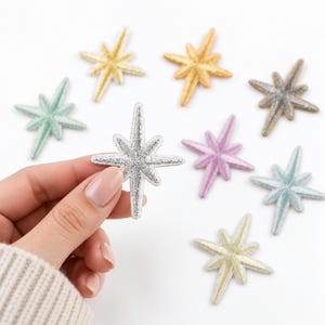 May include: A collection of eight embroidered star-shaped patches in various metallic and pastel colors, including silver, gold, mint green, lavender, and light blue. Each star features a pointed, multi-rayed design with a stitched border.
