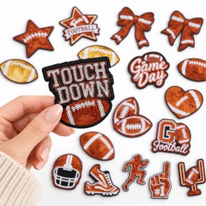 May include: A collection of orange and white football-themed embroidered patches featuring various designs including a football, a helmet, a goal post, a running player, a foam finger, bows, stars, and hearts, with one patch reading TOUCH DOWN.