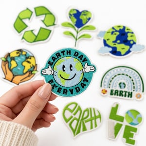May include: A collection of embroidered patches featuring environmental themes, including a circular patch with a smiling Earth character and the text EARTH DAY EVERYDAY, a green recycling symbol, a heart-shaped plant, and a rainbow with the word EARTH.