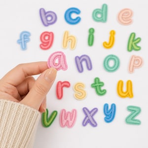 May include: A collection of lowercase alphabet letter patches in various pastel colors including pink, blue, green, yellow, and purple. Each letter features a stitched border and a soft, felt-like texture, arranged in a grid pattern on a white surface.