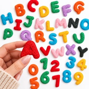 May include: A collection of colorful, textured, fuzzy alphabet letters and numbers scattered across a white surface. The letters and numbers are rendered in various bright shades including red, orange, yellow, green, blue, pink, purple, and black.