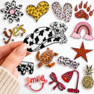 May include: A collection of embroidered iron-on patches featuring various designs including a cow print cowboy hat, a leopard print heart, a rainbow, a star, a pineapple, a turtle, a dalmatian, a jeep, and text reading yes, cow, and smile.