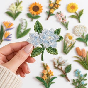 May include: A collection of embroidered flower patches featuring various botanical designs including sunflowers, lilies, and hydrangeas. One light blue flower patch with green leaves is held in the foreground, showcasing detailed stitching and vibrant colors.