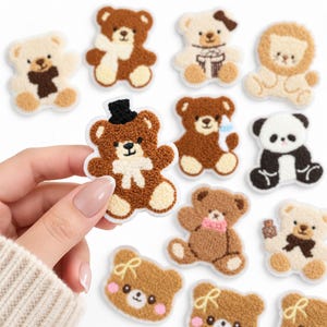 May include: A collection of small, textured, embroidered bear patches in various colors including brown, cream, and black and white. Each patch features a unique bear design, some with accessories like hats, scarves, or bows, arranged on a white surface.