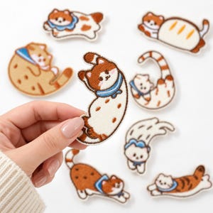 May include: A collection of embroidered cat patches featuring various brown, white, and orange feline designs in playful poses. Each patch depicts a small cat with a blue collar, showcasing detailed stitching and distinct shapes including curved and stretched forms.