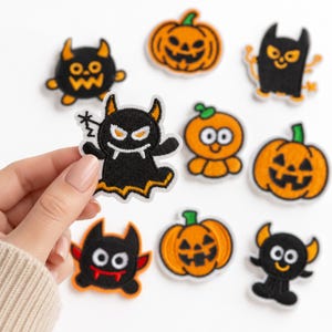 May include: A collection of embroidered Halloween-themed patches featuring various black monsters with orange accents and orange jack-o'-lantern pumpkins. The designs include round, horned creatures, smiling ghosts, and carved pumpkins with green stems.