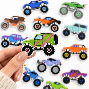 May include: A collection of embroidered monster truck patches in various colors including blue, green, orange, brown, and purple. Each patch features large black tires and detailed truck bodies. One lime green patch with pink stripes is held up.