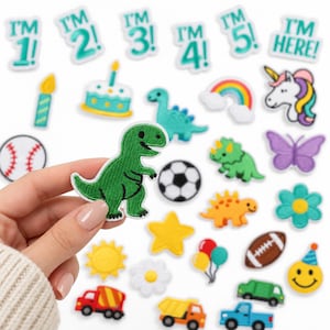 May include: A collection of embroidered iron-on patches featuring various colorful designs including dinosaurs, vehicles, sports balls, birthday themes, flowers, and text patches that read I'M 1!, I'M 2!, I'M 3!, I'M 4!, I'M 5!, and I'M HERE!.