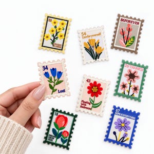 May include: A collection of seven rectangular embroidered patches shaped like postage stamps, each featuring a unique floral design, a number, and text. The patches display various colors including yellow, blue, red, pink, green, and purple flowers.
