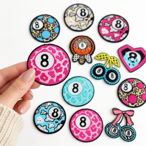 May include: A collection of embroidered patches featuring the number eight on various backgrounds, including leopard print, cow print, and cherry shapes. The patches are circular or heart-shaped with black borders, showcasing vibrant colors like pink, blue, and orange.