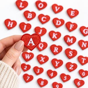 May include: A collection of small, red heart-shaped embroidered patches, each featuring a single white letter or number. One patch with the letter A is held up, while the others are scattered, displaying the full alphabet and digits zero through nine.