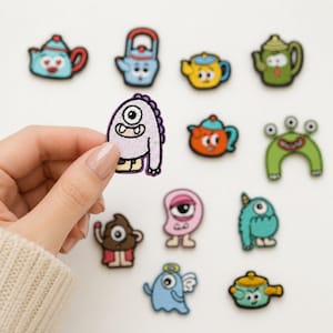 May include: A collection of embroidered patches featuring various cartoon characters and kitchenware items. The designs include colorful teapots, a green arch-shaped creature, a pink cyclops, a blue ghost with wings, and a light purple monster.