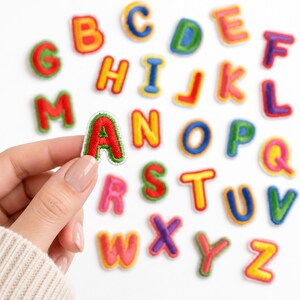 May include: A collection of colorful embroidered alphabet letter patches featuring various bright hues including red, yellow, blue, green, and purple. Each letter has a distinct border and is arranged in a scattered pattern on a white surface.