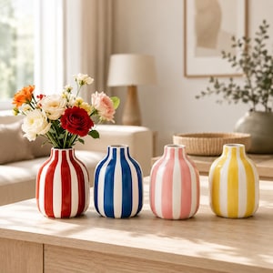 Può includere: Quattro piccoli vasi in ceramica dalla forma arrotondata con strisce verticali. I vasi sono di colore rosso, blu, rosa e giallo, ognuno con strisce bianche. Il vaso rosso contiene un bouquet di fiori colorati che include rose.
