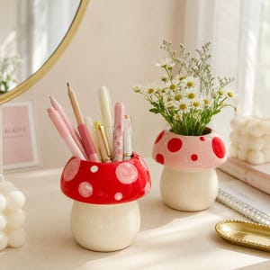 May include: Two ceramic mushroom-shaped containers. One is red with white spots, holding various pens and pencils. The other is light pink with red spots, containing a bouquet of small white daisy-like flowers with green stems and leaves.