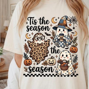 May include: A cream-colored t-shirt featuring a graphic print with the text 'Tis the season' and 'the season'. The design includes three ghosts, one with a leopard print, one wearing a blue plaid hat holding a coffee, and one with polka dots.