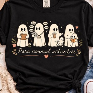 May include: A black short-sleeved t-shirt featuring a graphic of four ghosts engaged in various tasks with the text "Para normal activities" below them. The design includes small stars, hearts, and flowers in a light peach and white color palette.