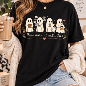 May include: A black short-sleeved t-shirt featuring a graphic of five white ghosts engaged in various tasks. The text "Para normal activities" is printed below the ghosts, accompanied by small floral accents and a heart symbol.