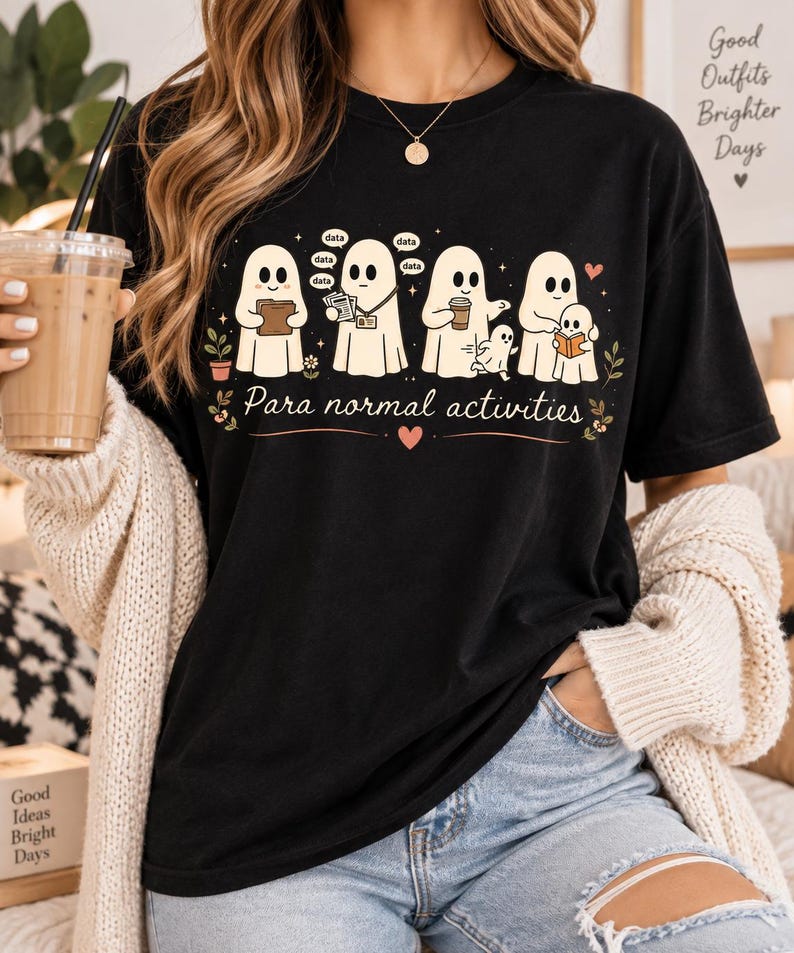May include: A black short-sleeved t-shirt featuring a graphic of five white ghosts engaged in various tasks. The text "Para normal activities" is printed below the ghosts, accompanied by small floral accents and a heart symbol.