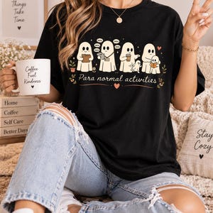 May include: A black t-shirt featuring a graphic of five white ghosts engaged in various tasks, with the text "Para normal activities" printed below them. The ghosts are shown reading, holding data, drinking coffee, and interacting with a smaller ghost.