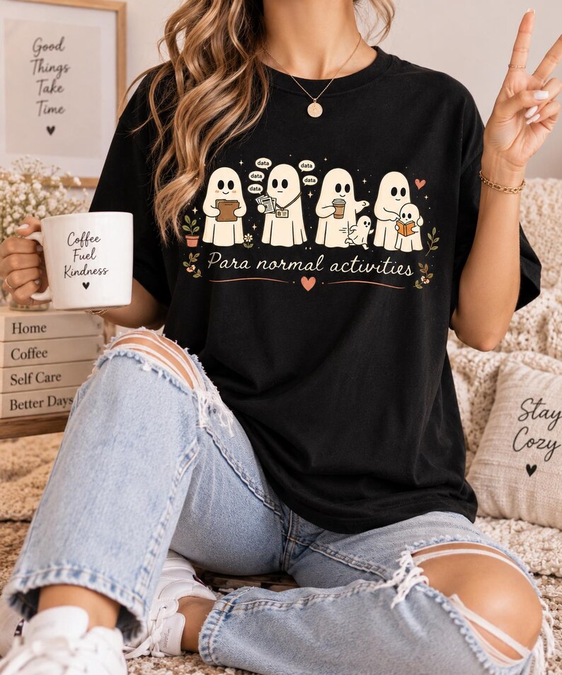 May include: A black t-shirt featuring a graphic of five white ghosts engaged in various tasks, with the text "Para normal activities" printed below them. The ghosts are shown reading, holding data, drinking coffee, and interacting with a smaller ghost.