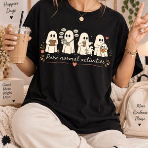 May include: A black t-shirt featuring a graphic of five white ghosts engaged in various office tasks, with the text "Para normal activities" printed below. The design includes small speech bubbles with the word "data" and decorative floral elements.