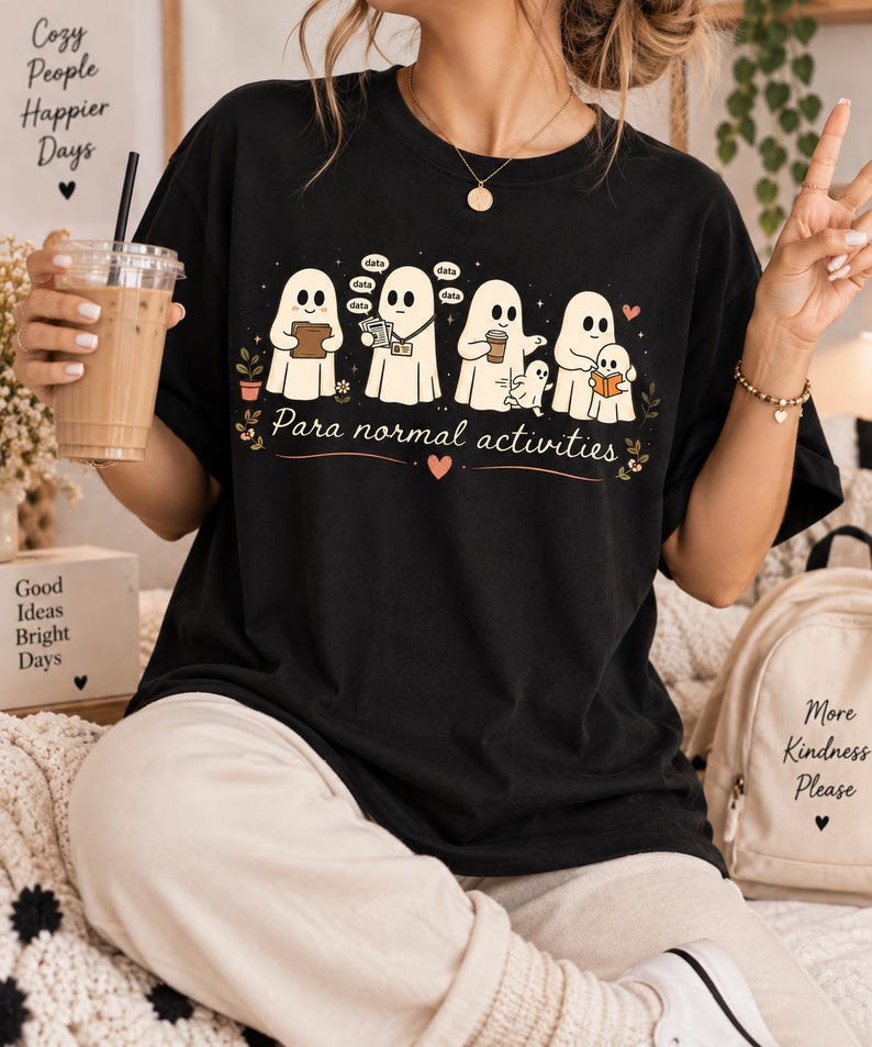 May include: A black t-shirt featuring a graphic of five white ghosts engaged in various office tasks, with the text "Para normal activities" printed below. The design includes small speech bubbles with the word "data" and decorative floral elements.