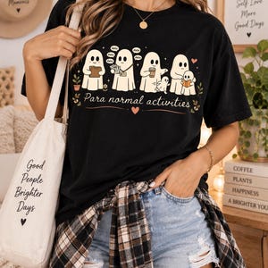 May include: A black t-shirt featuring a graphic of five white ghosts engaged in various tasks with the text "Para normal activities" printed below. The ghosts are shown holding a folder, papers, a coffee cup, and a book, with small speech bubbles.