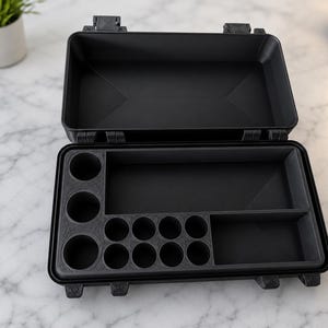 May include: A black rectangular storage case with a hinged lid and a custom interior insert. The insert features three large circular slots on the left, a long rectangular compartment, a smaller rectangular section, and eight small circular slots.