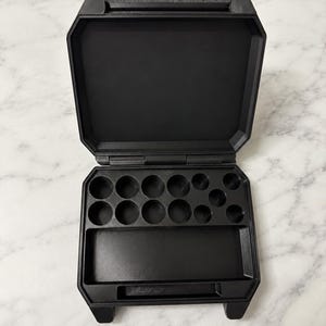 May include: A black, rectangular, hinged storage case with a matte finish. The interior features a large, flat rectangular compartment at the bottom and a series of circular indentations of varying sizes arranged in the upper section.