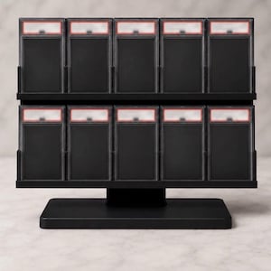 May include: A black tiered display stand holding ten rectangular trading card slabs. The stand features two horizontal rows, each containing five clear plastic cases with black inserts and red-bordered label areas at the top, set on a base.