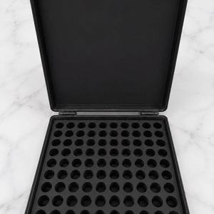May include: A black rectangular storage case with a hinged lid, featuring an interior grid of one hundred circular cutouts arranged in a ten-by-ten pattern. The case is constructed from a matte black material and includes two top latches.
