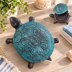 May include: Two turtle-shaped trinket boxes made of dark metal with a distressed teal finish on the shells. The larger turtle is positioned in the foreground, while the smaller turtle is shown with its shell lid removed to reveal a hollow interior.