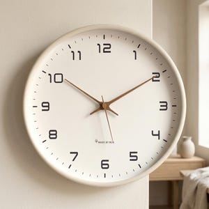 Puede incluir: Un reloj de pared redondo con marco y esfera de color crema. Presenta números arábigos negros para las horas, manecillas de metal marrón fino y el texto MADE BY MJK impreso en letras negras pequeñas cerca de la parte inferior central.