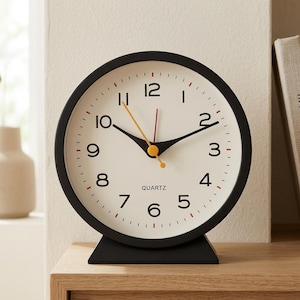 Può includere: Un orologio da tavolo al quarzo rotondo con cornice e base nero opaco. Il quadrante è bianco sporco con numeri arabi neri da 1 a 12. Presenta lancette nere per ore e minuti, una lancetta dei secondi arancione sottile e una linea rossa.
