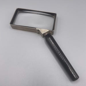 May include: A rectangular magnifying glass featuring a clear glass lens encased in a silver-toned metal frame. The handle is cylindrical, smooth, and black, connected to the frame by a small, triangular silver-toned metal joint piece.