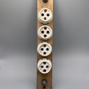 Op de afbeelding: Een verticaal rechthoekig houten bord met vier ronde witte keramische stopcontacten in een kolom. Elk stopcontact heeft drie kleine gaatjes. Aan zowel de boven- als onderkant van het houten paneel bevindt zich een metalen bout.