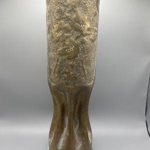 May include: A tall, cylindrical brass vase featuring a scalloped top edge and a textured surface. The exterior is decorated with an embossed floral pattern of flowers and stems, set against a stippled background, with a flared, fluted base.