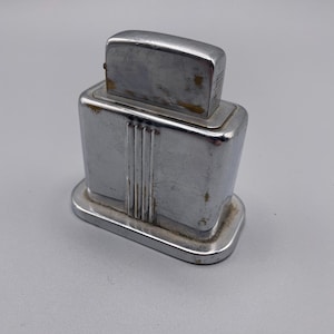 May include: A vintage chrome-plated table lighter with a rectangular base and a vertical ribbed design on the front. The lighter features a smooth, polished metallic finish with visible signs of wear and oxidation along the edges and corners.