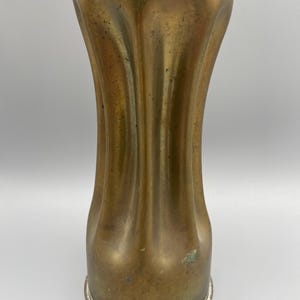 May include: A tall, cylindrical brass vase featuring a vertical fluted design with deep, rounded grooves. The metal surface displays a weathered, aged patina with subtle variations in color, including hints of oxidation and dark spots.