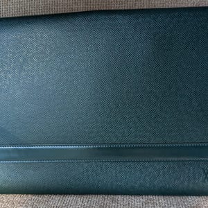 May include: A dark green rectangular document holder featuring a textured cross-hatch finish. The item includes a horizontal band across the lower section and a small embossed LV logo in the bottom right corner, presenting a structured design.