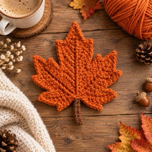 May include: A crocheted maple leaf coaster in a vibrant burnt orange color with a dark brown stem. The leaf features a textured, hand-stitched design with defined veins and pointed lobes, crafted from yarn to resemble a fallen autumn foliage piece.