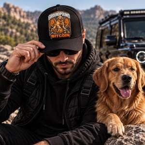 May include: A black baseball cap featuring a graphic print of a golden retriever sitting on a pile of gold coins with the text GROUNDPOUND BITCOIN. The design includes orange Bitcoin symbols on either side of the dog against a dark background.