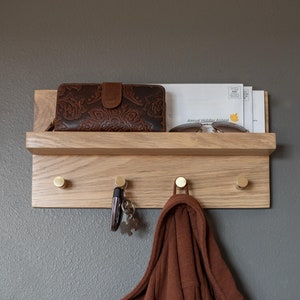 May include: A wooden wall mounted shelf with hooks and a small shelf on top. The shelf has a brown leather wallet, sunglasses, and some mail on it. There is a brown sweater hanging on one of the hooks and keys on another hook.