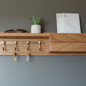 May include: A wooden key rack with a shelf and a mail slot. The key rack has five hooks and is mounted on a wall. There are two keys hanging on the hooks and a pair of sunglasses on the shelf. A small potted plant is on the shelf.