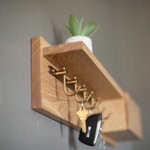 May include: A wooden wall-mounted shelf with hooks for hanging keys and a small shelf for displaying items. The shelf is made of light wood and has a natural finish. There are five hooks on the shelf, and a small potted plant is sitting on the shelf.