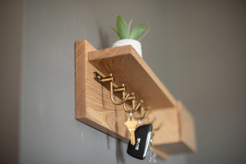 May include: A wooden wall-mounted shelf with hooks for hanging keys and a small shelf for displaying items. The shelf is made of light wood and has a natural finish. There are five hooks on the shelf, and a small potted plant is sitting on the shelf.