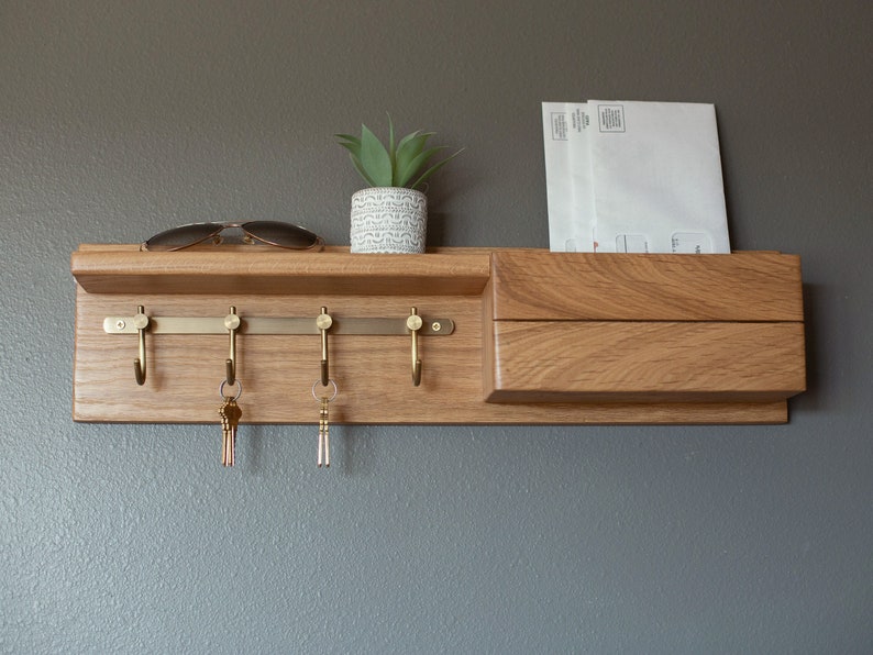 May include: A wooden key rack with a shelf and a mail slot. The key rack has five hooks and is mounted on a wall. There are two keys hanging on the hooks and a pair of sunglasses on the shelf. A small potted plant is on the shelf.